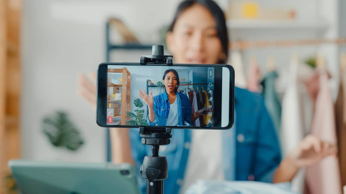 young asia lady fashion designer using mobile phone receiving purchase order and showing clothes recording video live streaming online at shop.